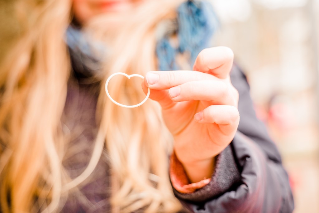 A person holding a white heart-shaped object with long, wavy hair and wearing a jacket and scarf.