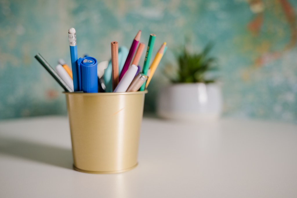 A gold container filled with assorted pens and markers, placed on a table with a blurred background featuring a small potted plant.