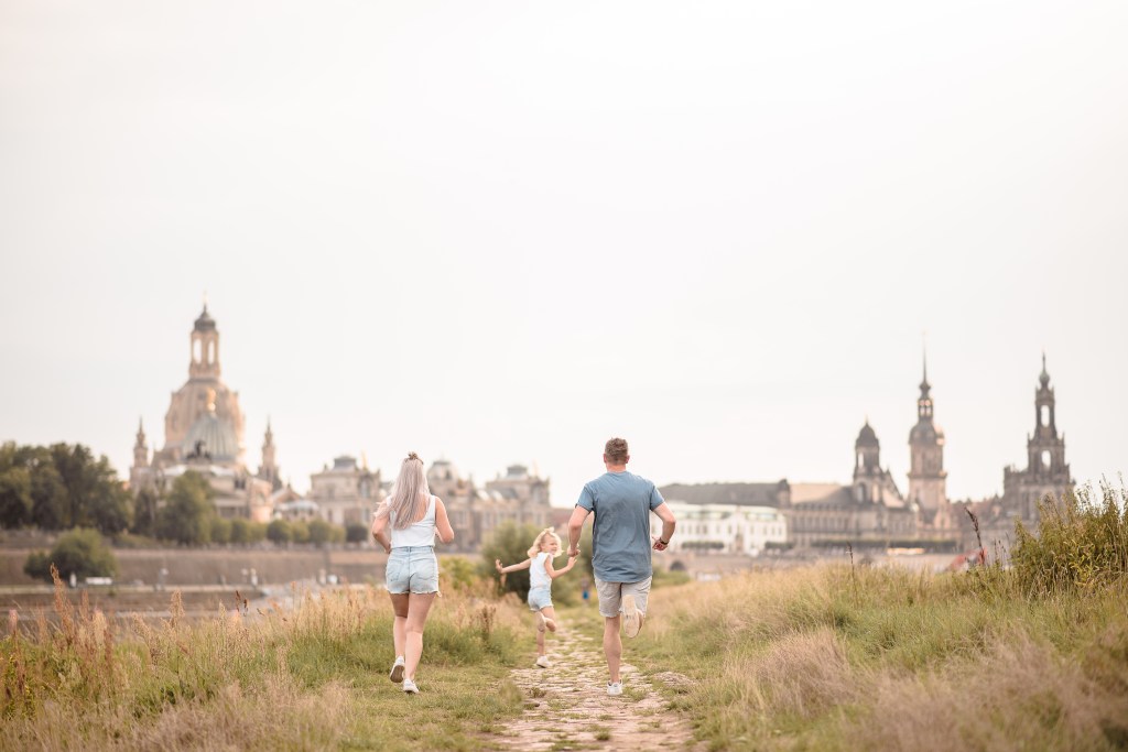 Eine Familie mit einem Kind, das auf einem Weg in der Natur läuft, während die Stadtansicht von Dresden im Hintergrund sichtbar ist.