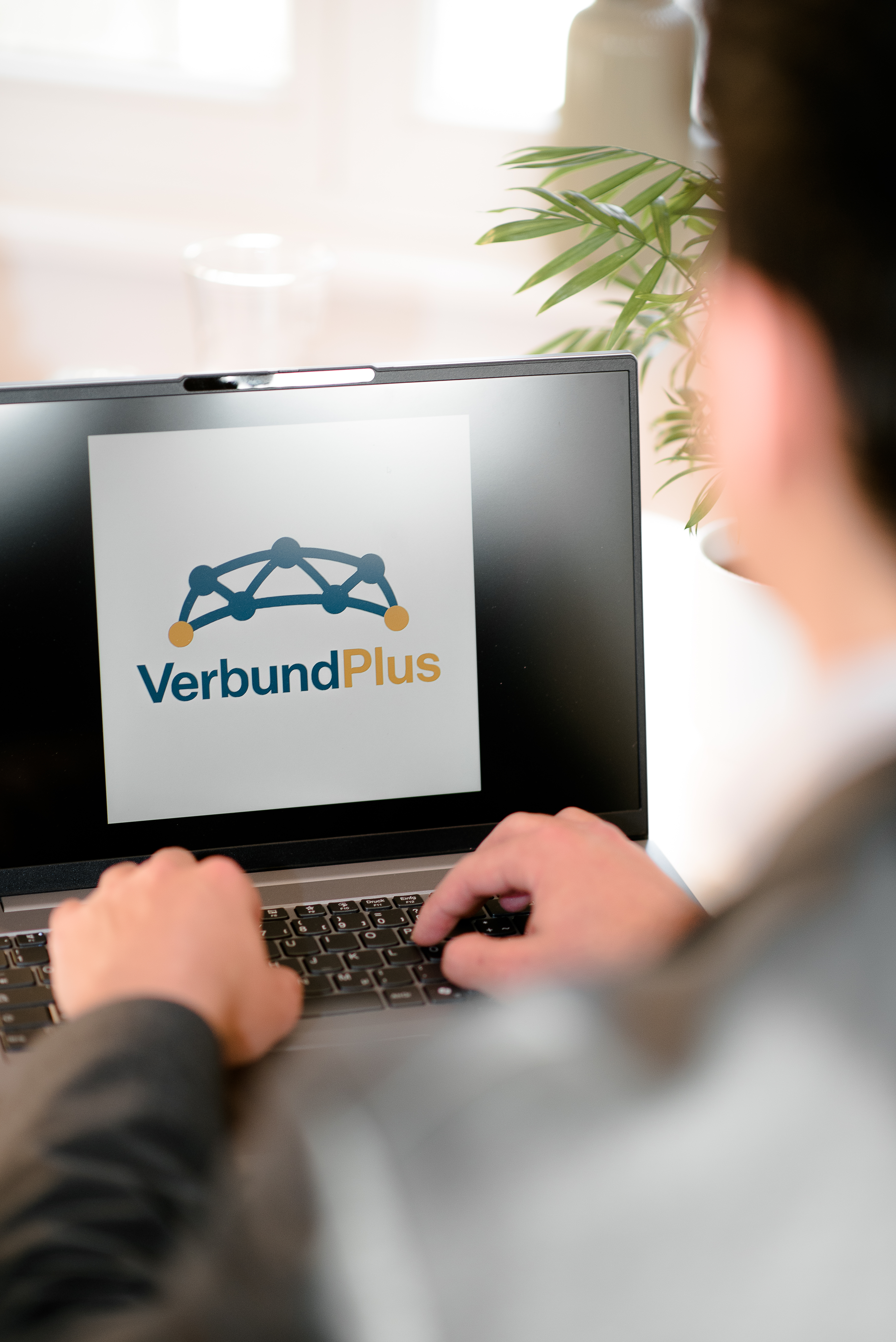 Eine Person, die am Laptop sitzt und das Logo von VerbundPlus auf dem Bildschirm betrachtet.