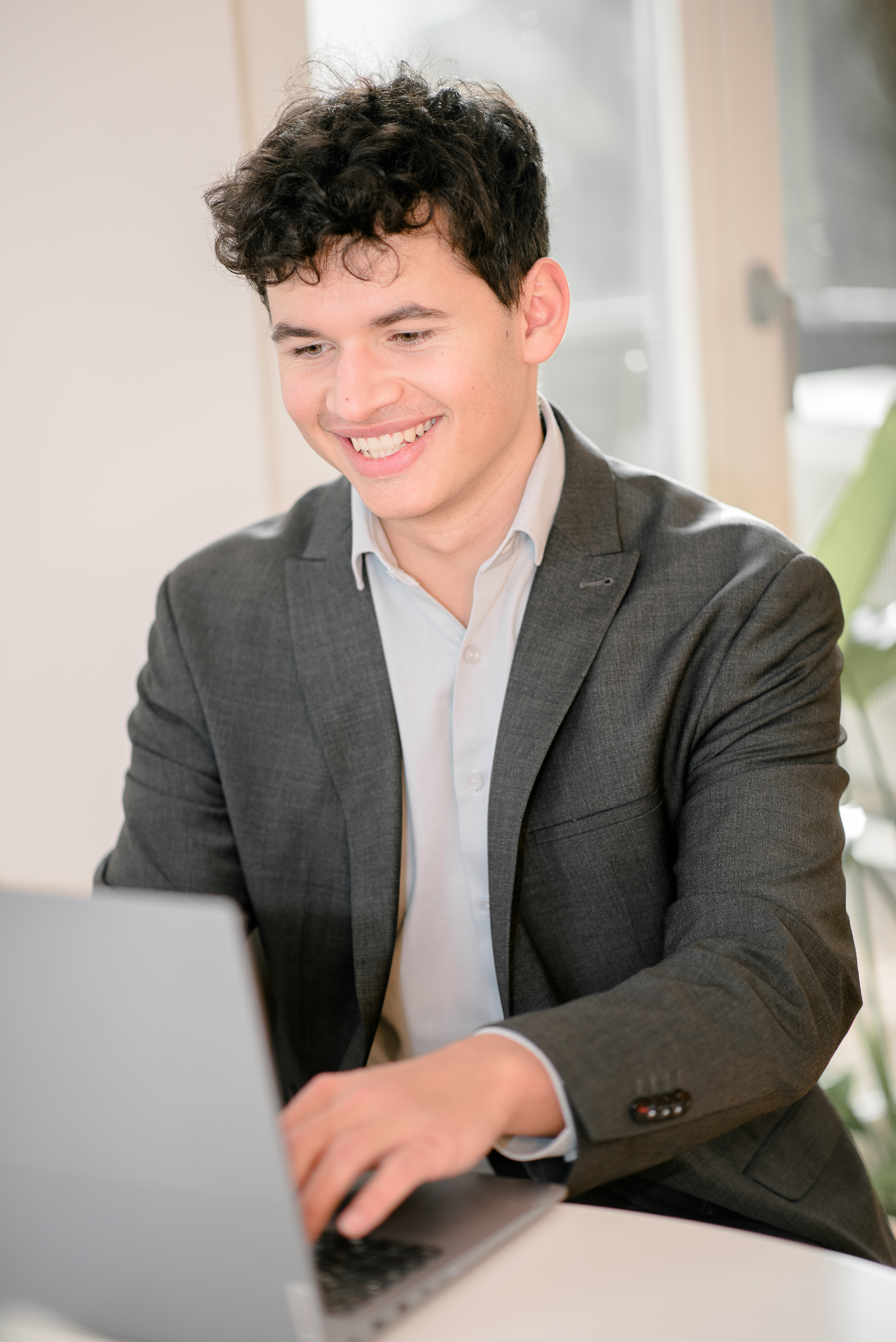 Unser Geschäftsführer, Jonathan Rößner, in einem grauen Anzug sitzt an einem unserer Tische und arbeitet am Laptop. Er lächelt, während er auf den Bildschirm schaut.