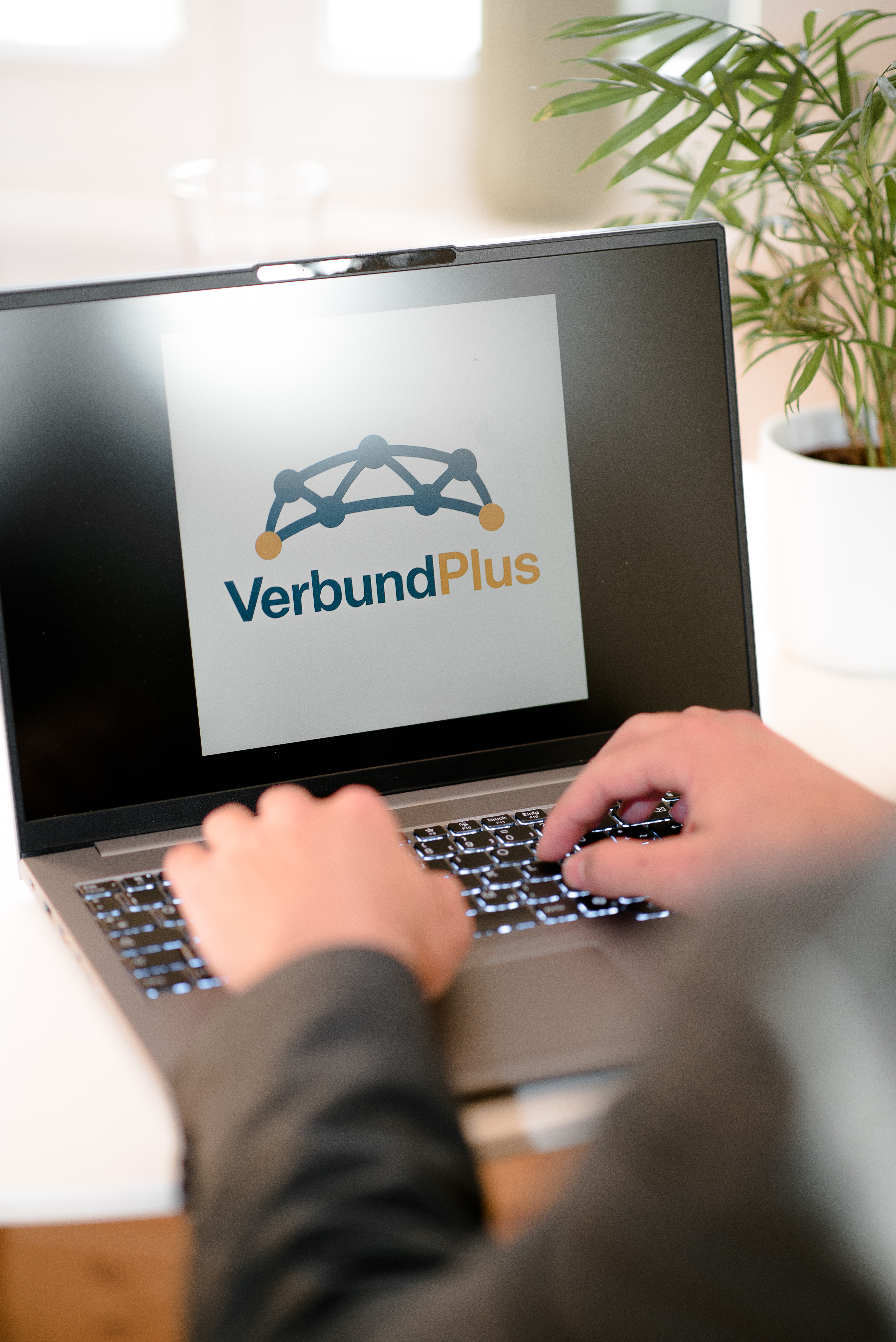 Eine Person sitzt vor einem Laptop und tippt, während das Logo von VerbundPlus auf dem Bildschirm angezeigt wird. Im Hintergrund sind eine Pflanze und eine helle Umgebung sichtbar.