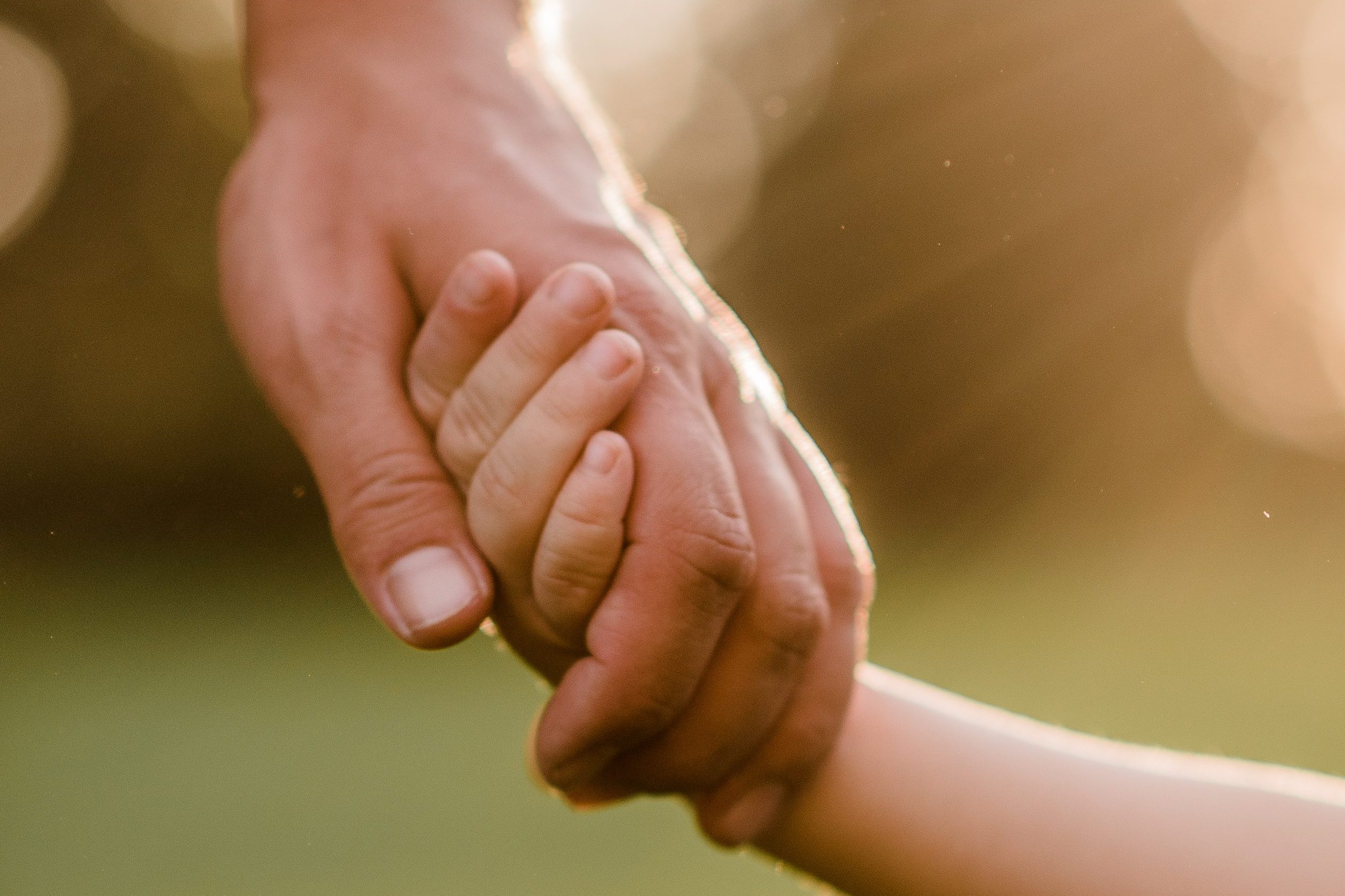Eine große Hand hält eine kleine Hand, umgeben von einem verschwommenen, warmen Licht im Hintergrund.